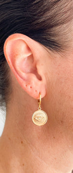 Load image into Gallery viewer, Evil Eye coin shaped pendant hanging from a gold filled huggie earring. The Evil Eye is a strong protection symbol or talisman.
