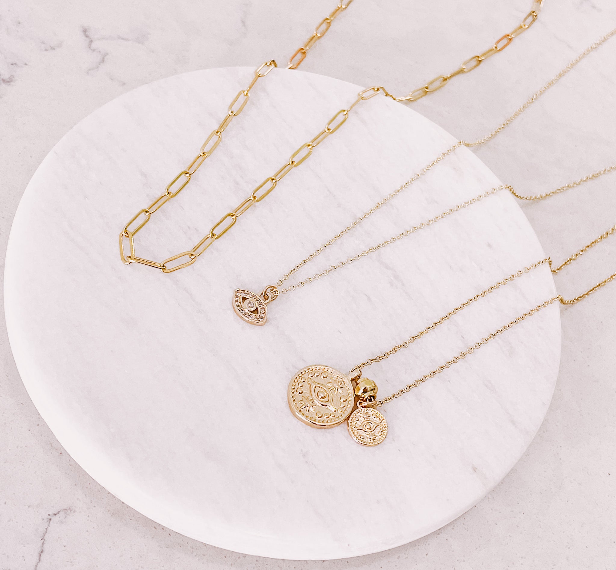 Flat lay shot of AW Boutique's Paperclip Chain Necklace, laying next to the Mini Crystal Evil Eye Necklace and the Dual Evil Eye Coin Necklace.